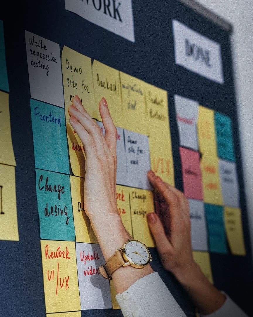 Hands moving handwritten sticky notes between columns on a physical task board.