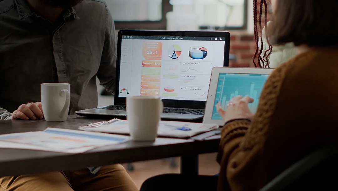 Two colleagues review colourful analytics dashboards on laptops across a meeting table.