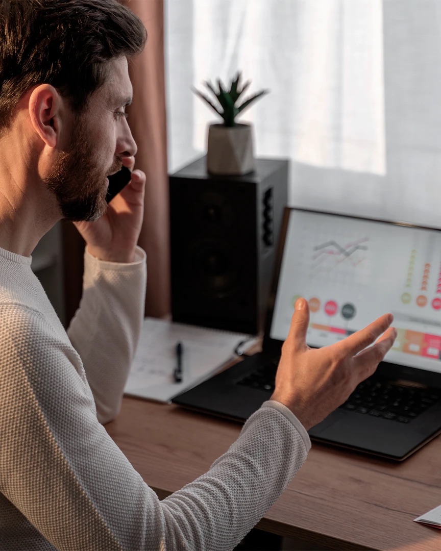 A man on the phone gestures towards a laptop showing performance charts and dashboards.